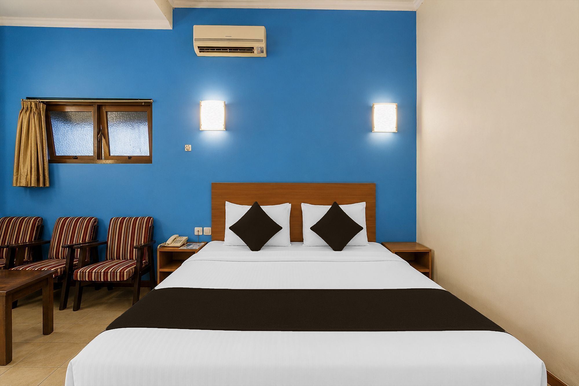 A hotel room with a large bed, two bedside lamps, a window with curtains, a desk with a telephone, and striped chairs.