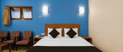 A hotel room with a large bed, two bedside lamps, a window with curtains, a desk with a telephone, and striped chairs.