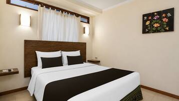 A hotel room with a bed, a nightstand, a wall-mounted light, a window with curtains, and a floral painting.