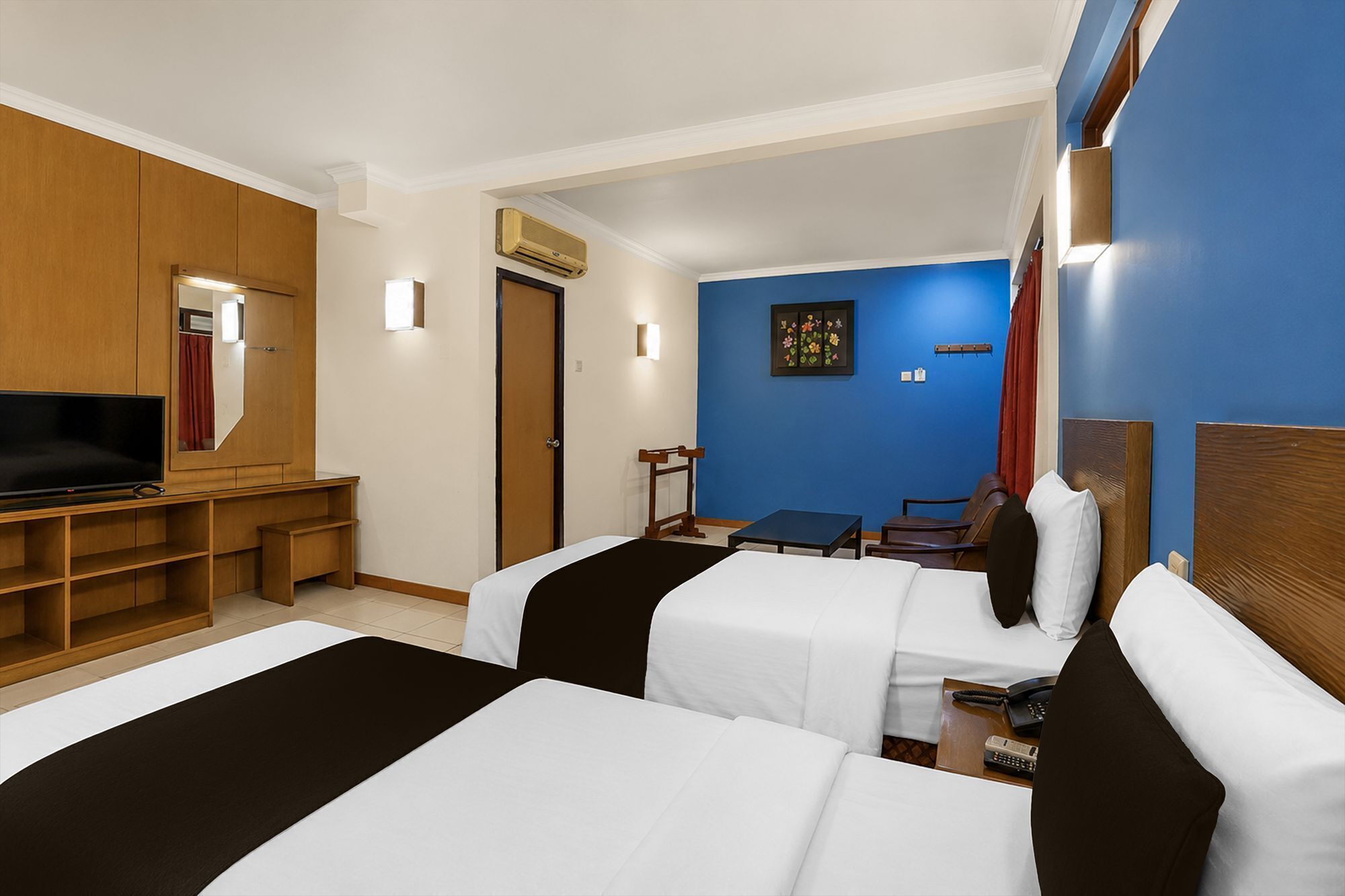 A hotel room with two beds, a TV, a desk, and a blue accent wall.