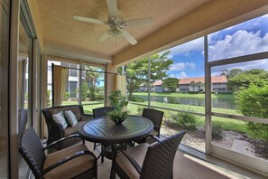 Eine überdachte Terrasse mit einem runden Tisch und Stühlen, große Fenster mit Blick auf einen See und Gebäude.