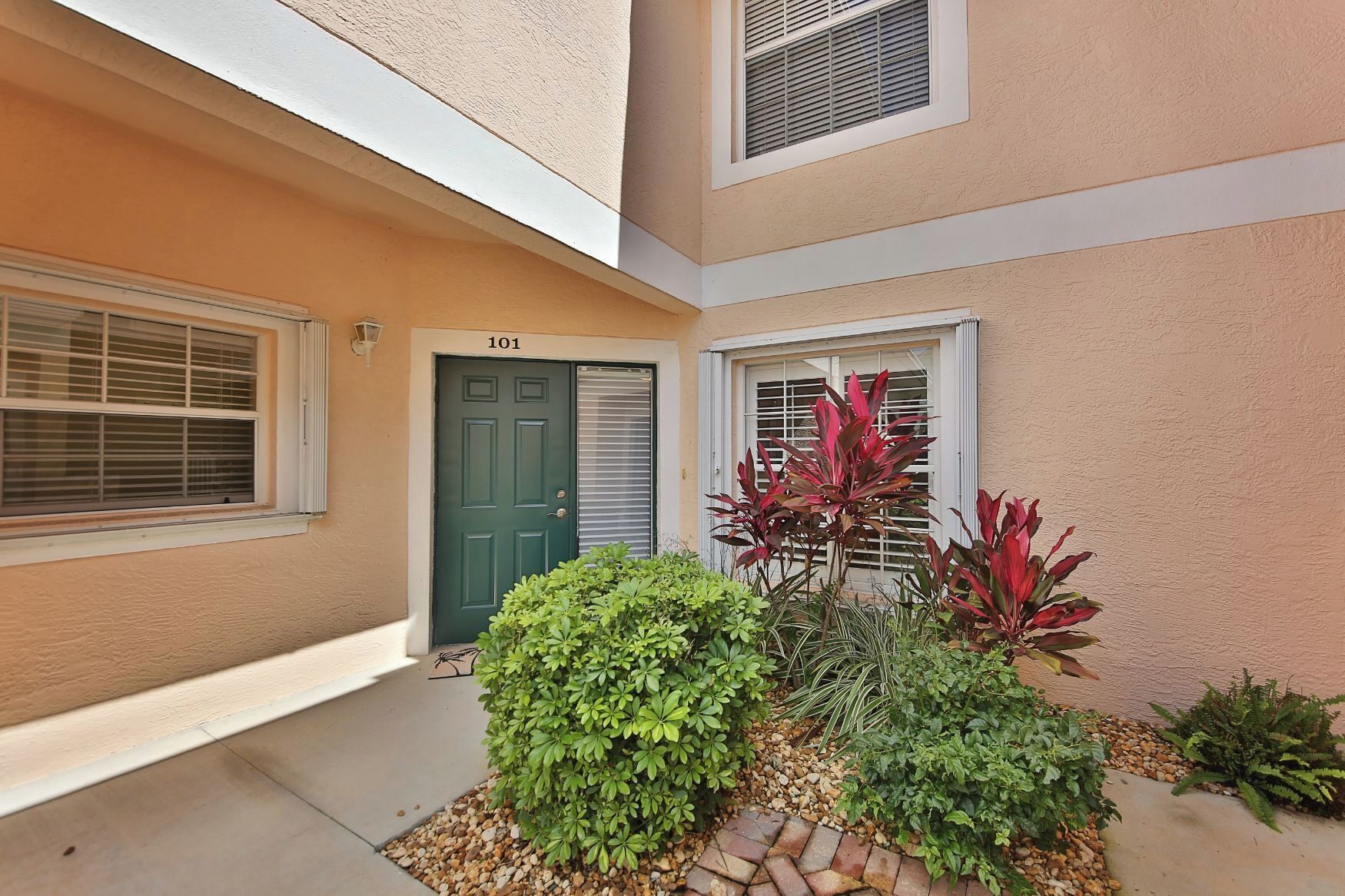 A residential building with a green door, a window with blinds, and a small garden with various plants.