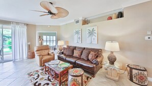 A living room with a brown leather sofa, a glass coffee table, a patterned rug, a ceiling fan, and a view of the outdoors through a glass door. - First Floor With a View of the Lake (Naples)
