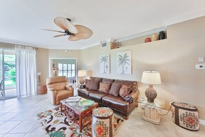 Ein Wohnzimmer mit braunem Ledersofa, einem Glastisch, einem gemusterten Teppich, einem Deckenventilator und Blick ins Freie durch eine Glastür.