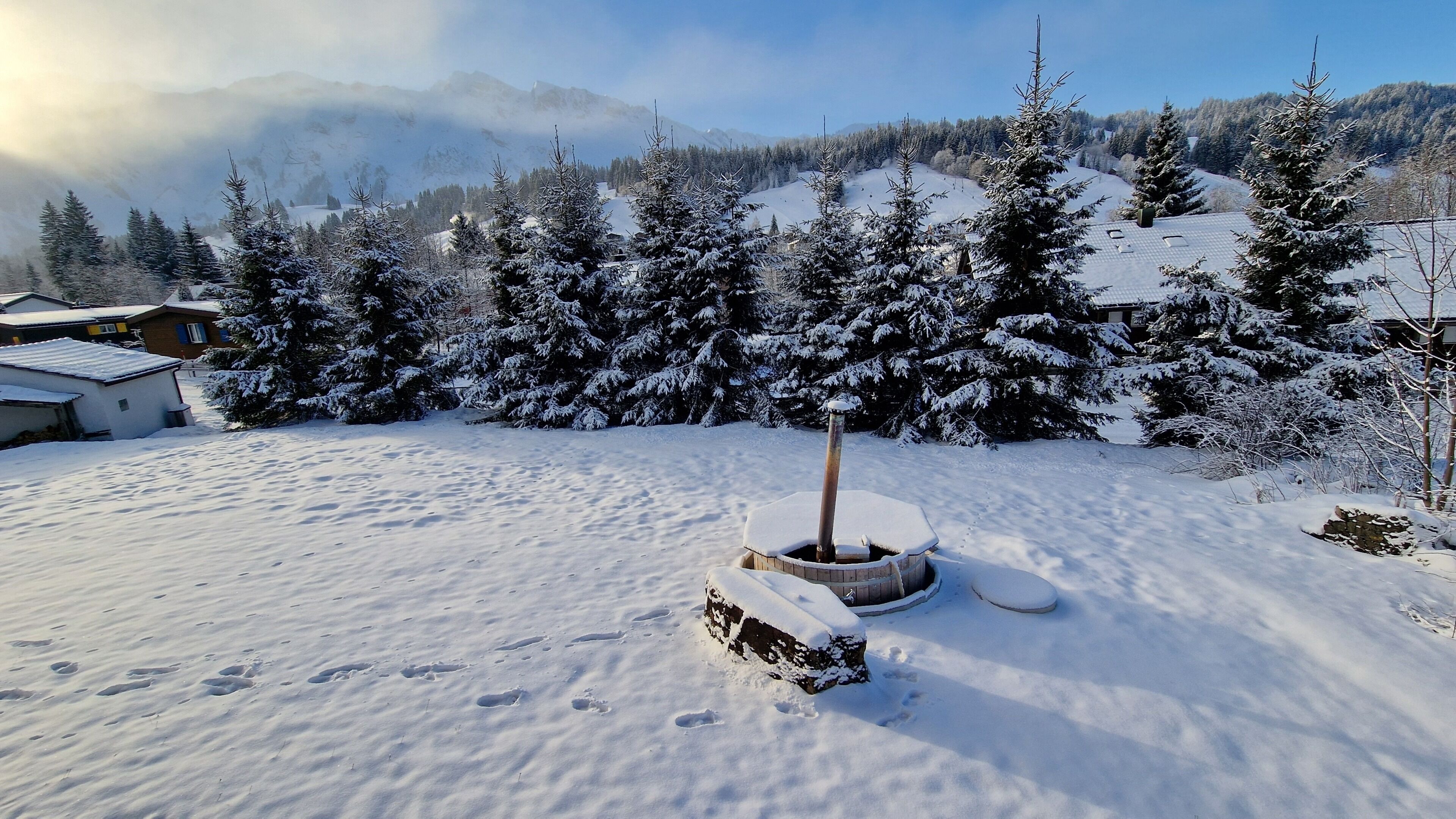 Eine verschneite Landschaft mit einer Holzhütte, möglicherweise eine Sauna, umgeben von schneebedeckten Bäumen und Häusern.