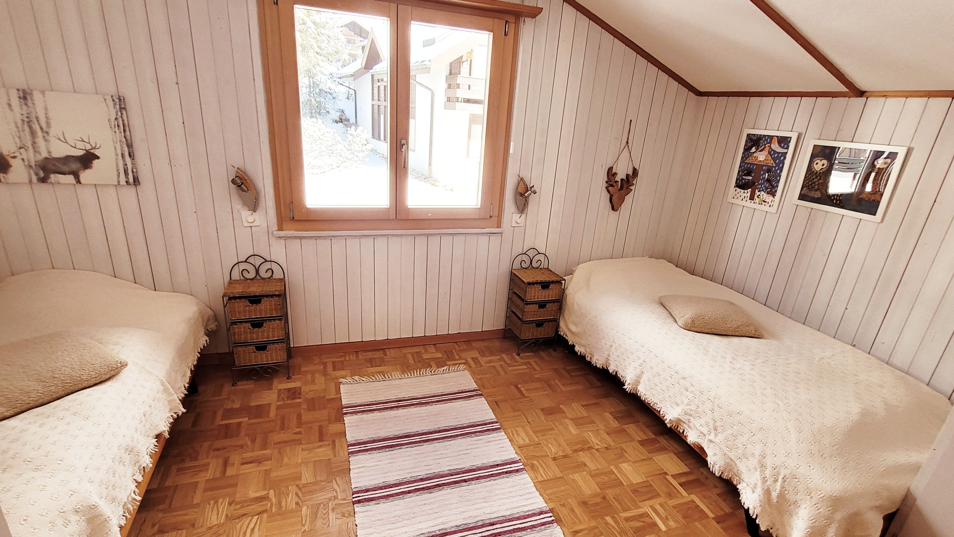 Ein Zimmer mit zwei Einzelbetten, einem Fenster, Holzwänden und einem Fischgrätparkett.