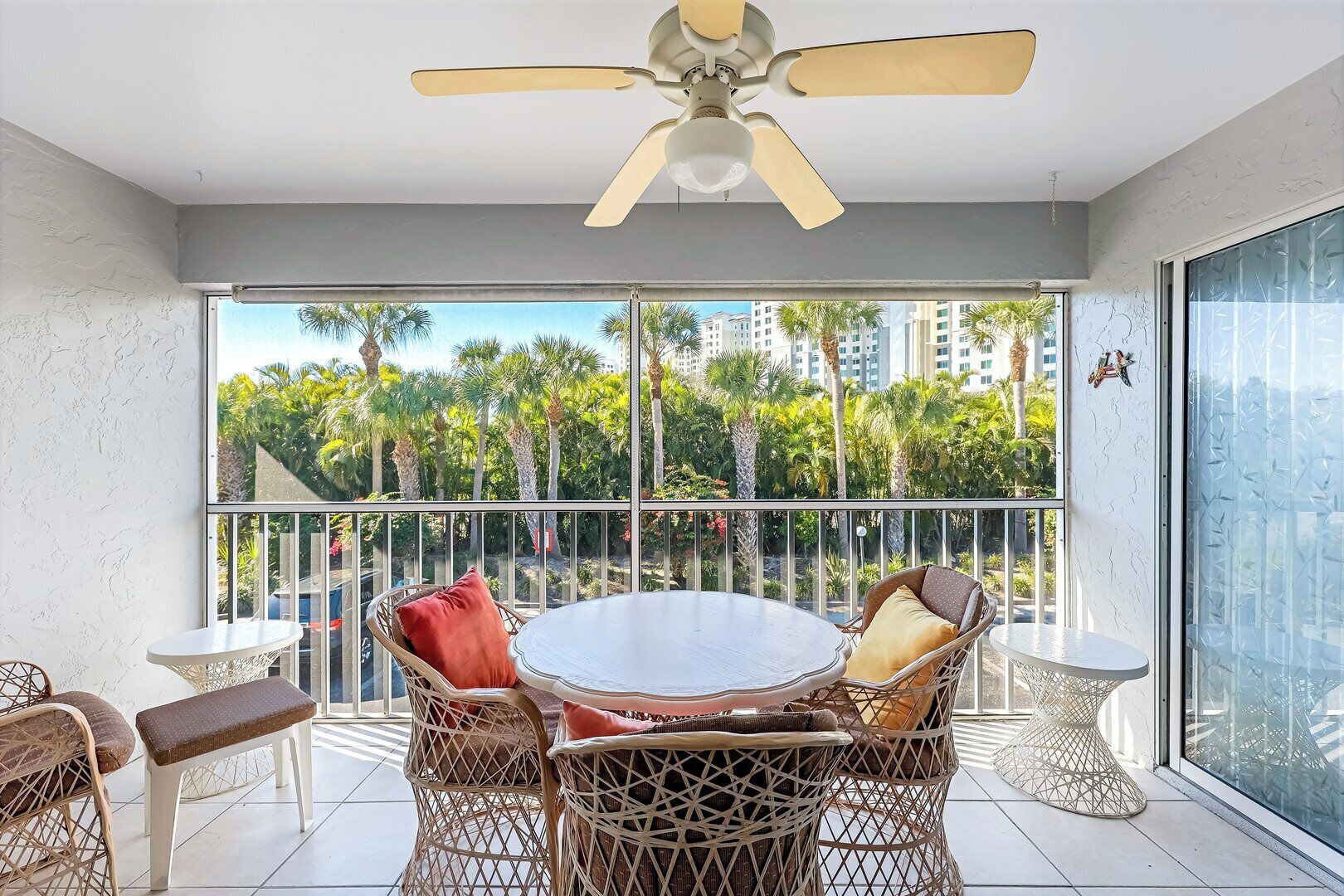 A balcony with a round table and wicker chairs, a ceiling fan, and a view of palm trees and buildings.