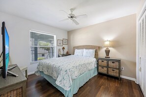A bedroom with a bed, a nightstand, a lamp, a ceiling fan, a television, and a window with blinds. - Walk to the Beach from Vanderbilt Villas (Naples)