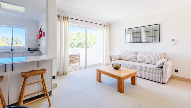 A modern living room with a sofa, a coffee table, and a kitchen area with a window. - Jardins do Golfe Top Floor Retreat (Loulé)