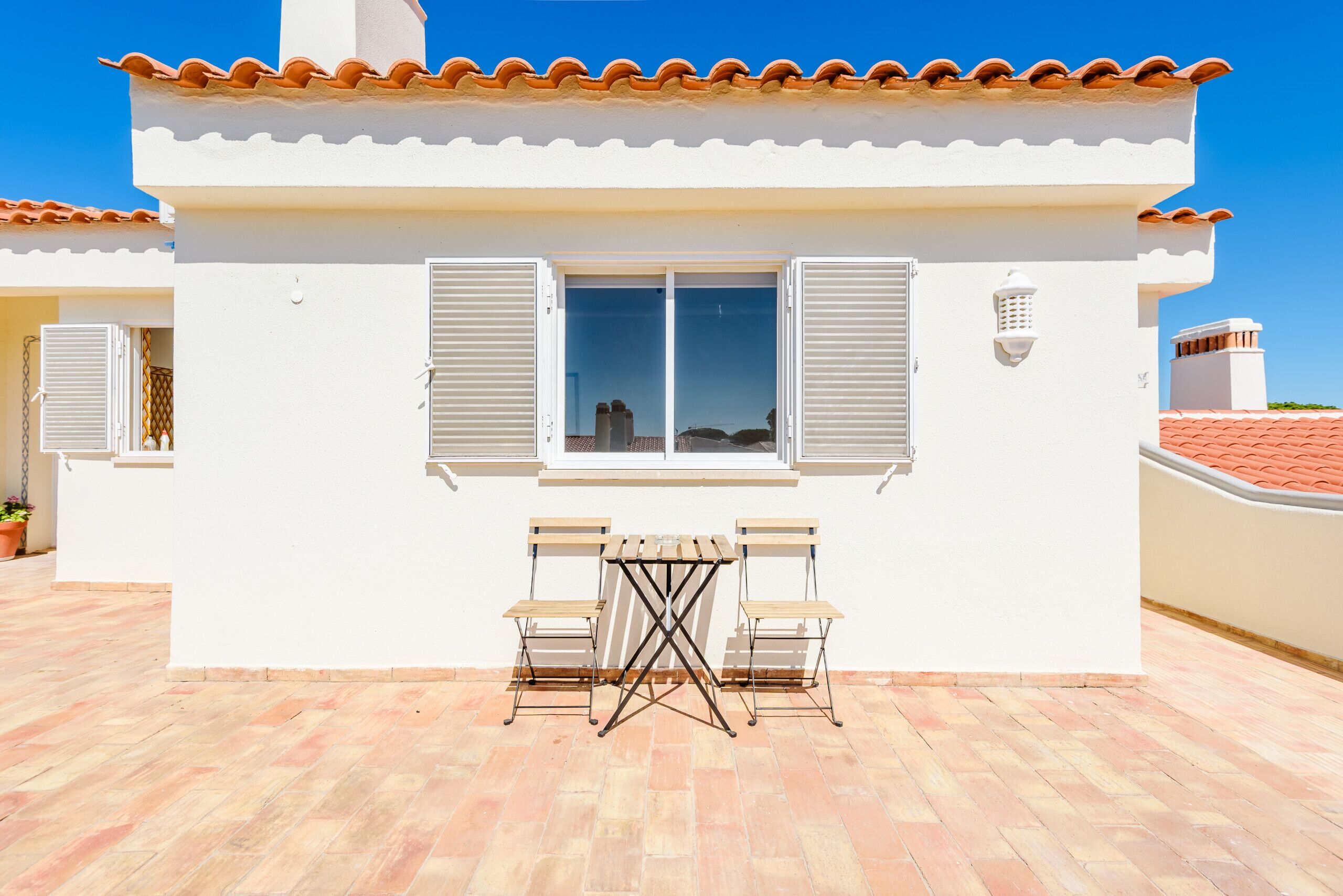 Un edificio blanco con techo de tejas, una ventana con contraventanas y un patio con dos sillas y una mesa.