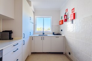A compact kitchen with white cabinets, a window, and emergency equipment on the wall. - Jardins do Golfe Top Floor Retreat (Loulé)