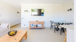 A modern living room with a sofa, a wooden table, a dining area with a glass table and chairs, and a wall-mounted TV. - Jardins do Golfe Top Floor Retreat (Loulé)