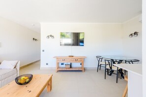 A modern living room with a sofa, a wooden table, a dining area with a glass table and chairs, and a wall-mounted TV. - Jardins do Golfe Top Floor Retreat (Loulé)
