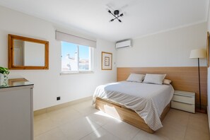 A bedroom with a bed, a bedside table, a lamp, a window, and an air conditioning unit. - Jardins do Golfe Top Floor Retreat (Vale do Lobo)