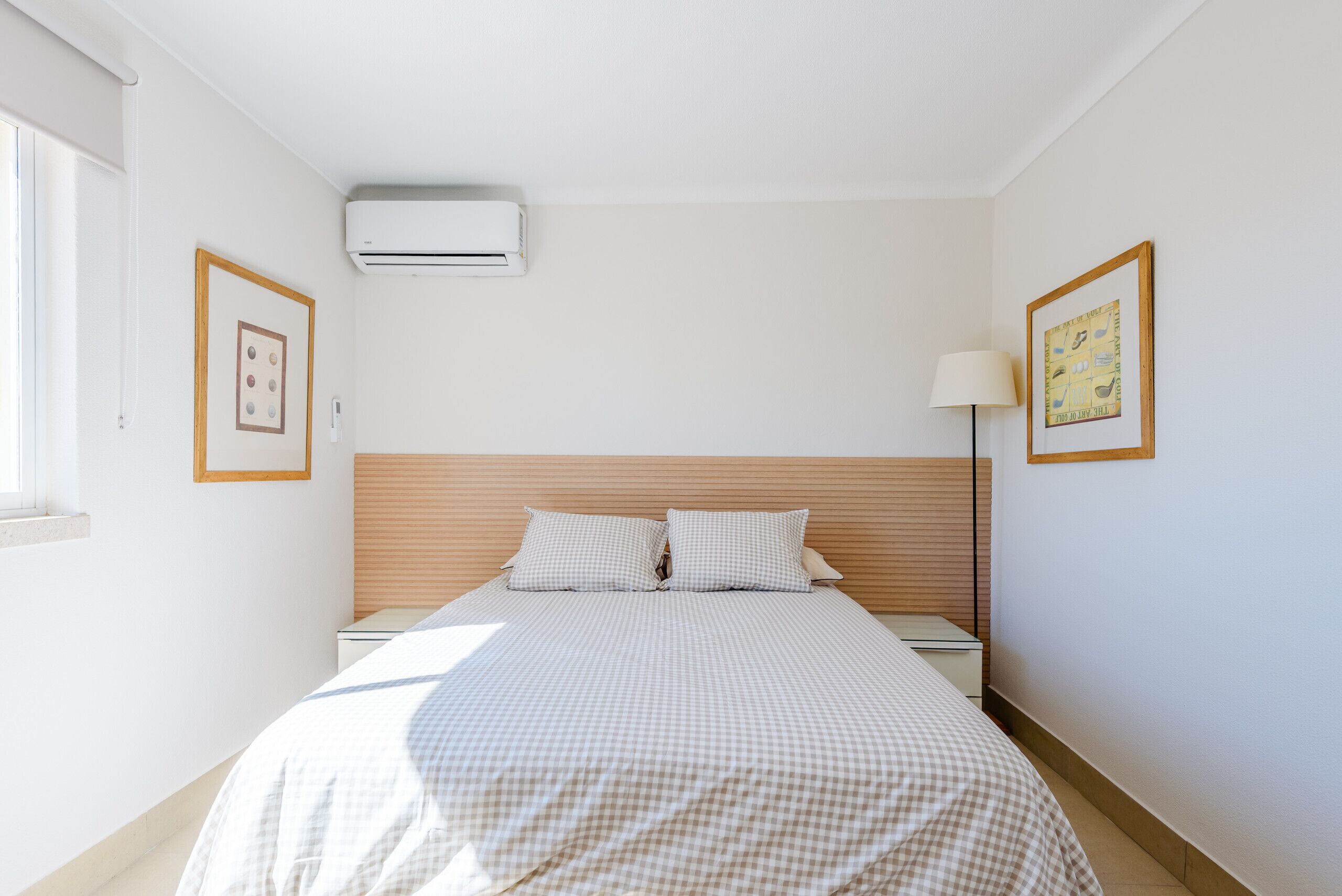 A neatly made bed with checkered bedding, a wooden headboard, and a wall-mounted air conditioner.