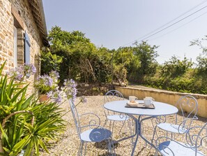 Ein Steinhaus mit einer Terrasse, auf der ein runder Tisch und Metallstühle stehen, umgeben von Grünpflanzen und einer Steinsäule.