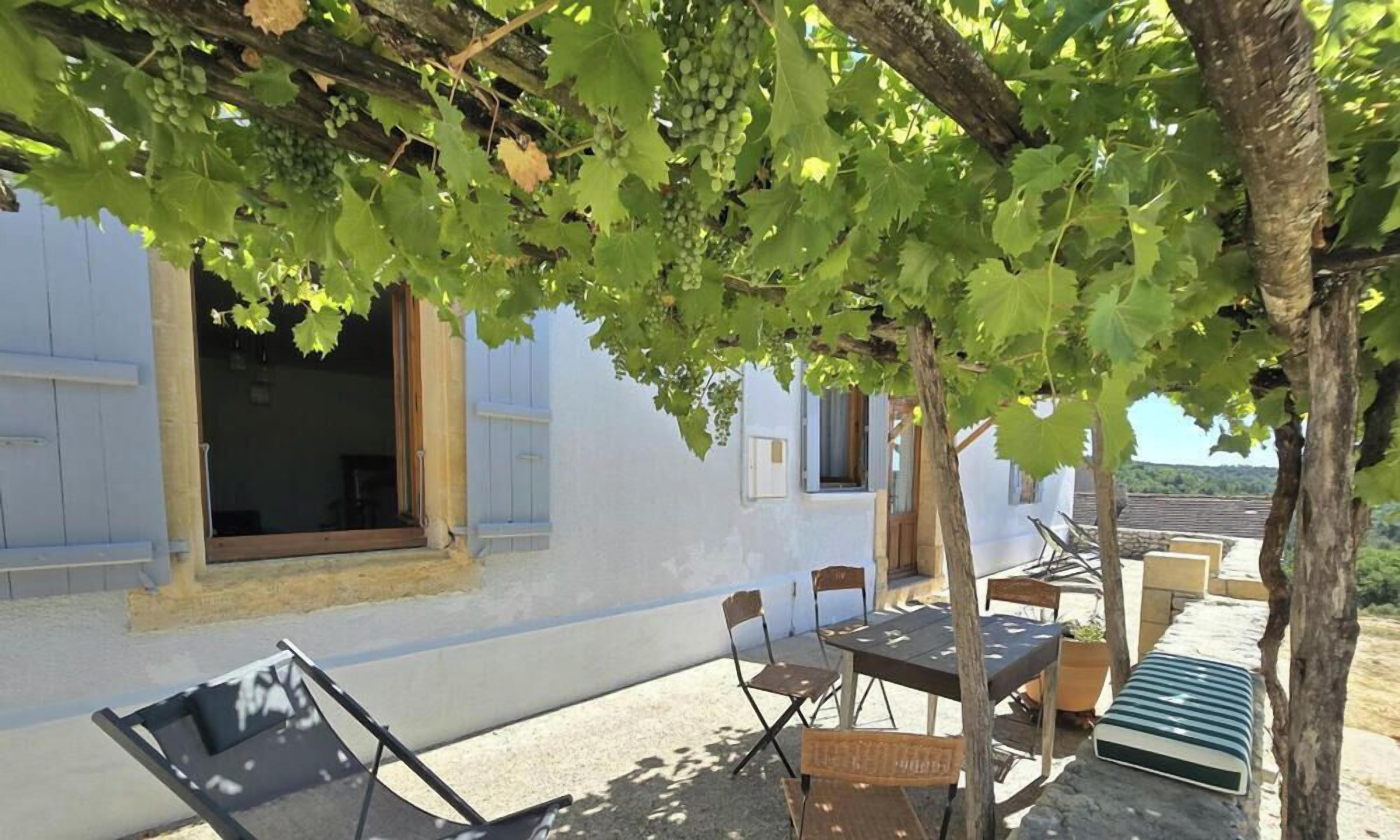 Un bâtiment à l’extérieur blanc, une table avec des chaises et une pergola couverte de vignes.