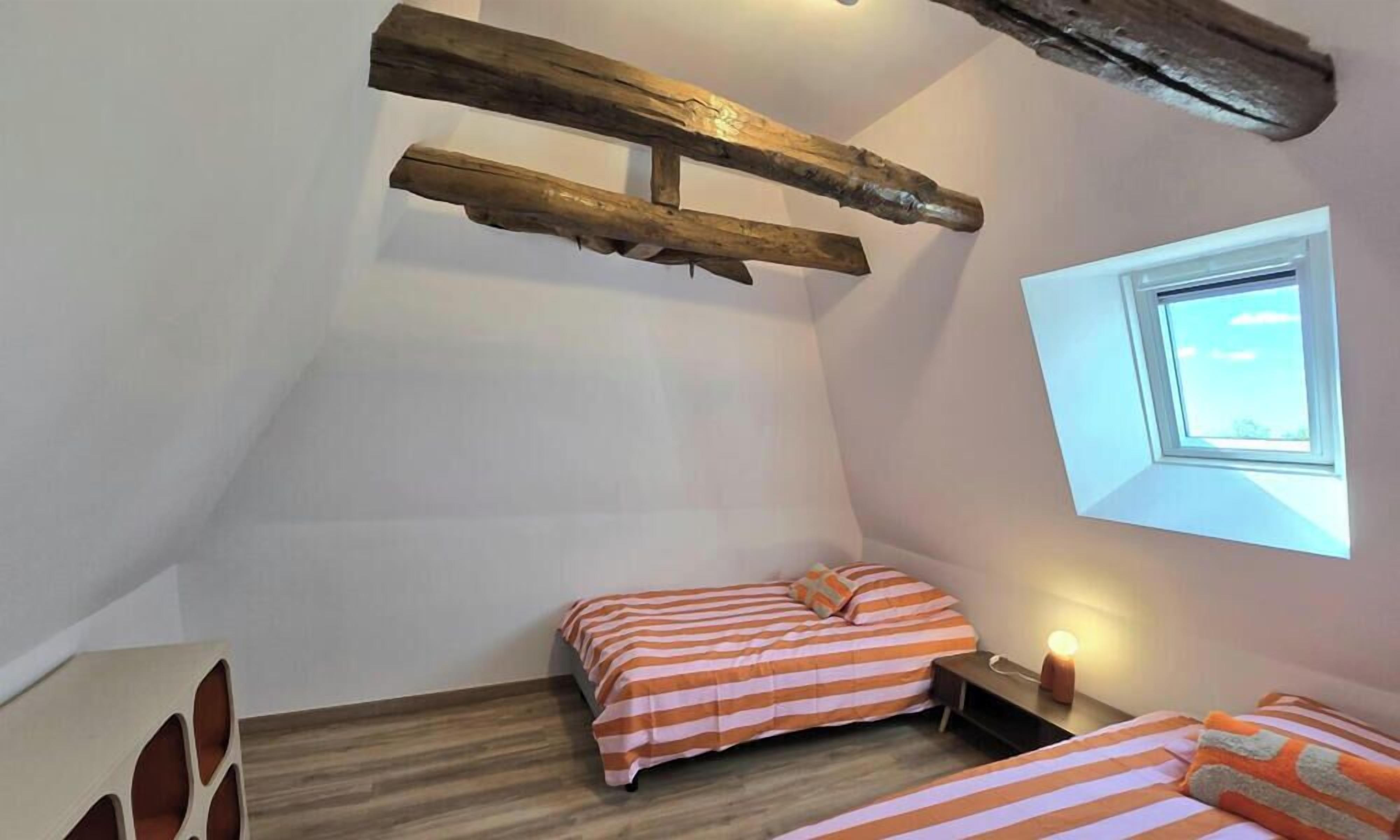 A small, sloped room with two beds, a wooden beam ceiling, a window, and a bedside lamp.
