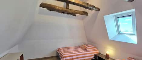 Ein kleiner Raum mit schrägem Dach, zwei Betten, Holzbalkendecke, einem Fenster und einer Nachttischlampe.