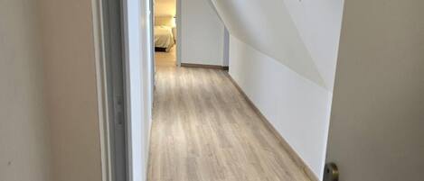 A hallway with wooden flooring and white walls, leading to a room with a bed.