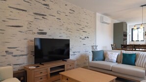 Una sala de estar moderna con un televisor de pantalla plana, muebles de madera, un sofá con cojines y una zona de comedor con sillas.