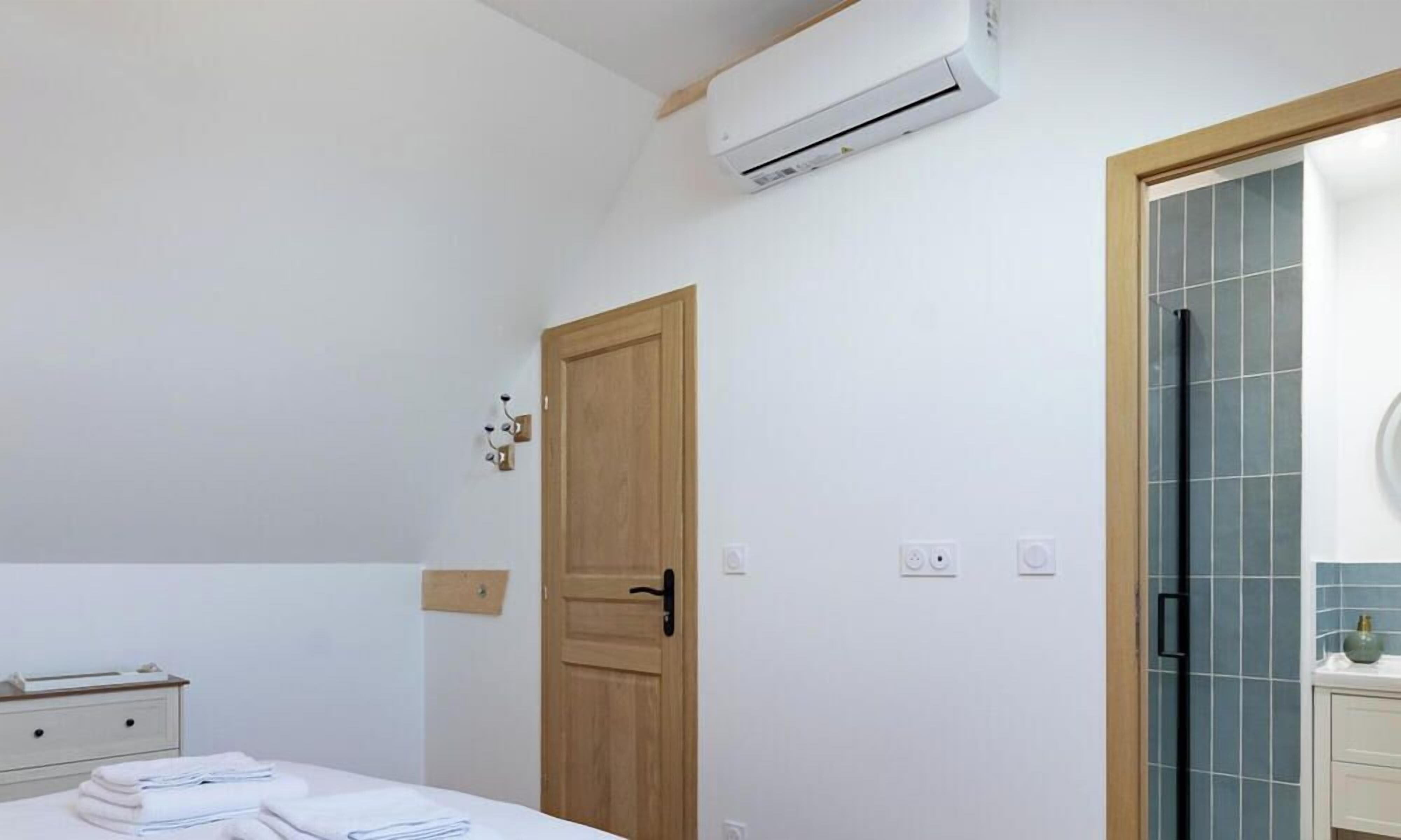 A hotel room with a bed, a wooden door, an air conditioning unit, a bathroom with a tiled wall, and a small bedside table with towels.