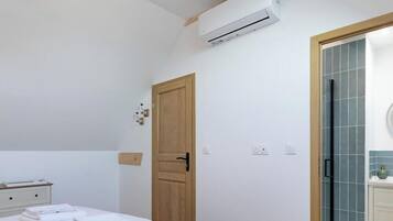 A hotel room with a bed, a wooden door, an air conditioning unit, a bathroom with a tiled wall, and a small bedside table with towels.