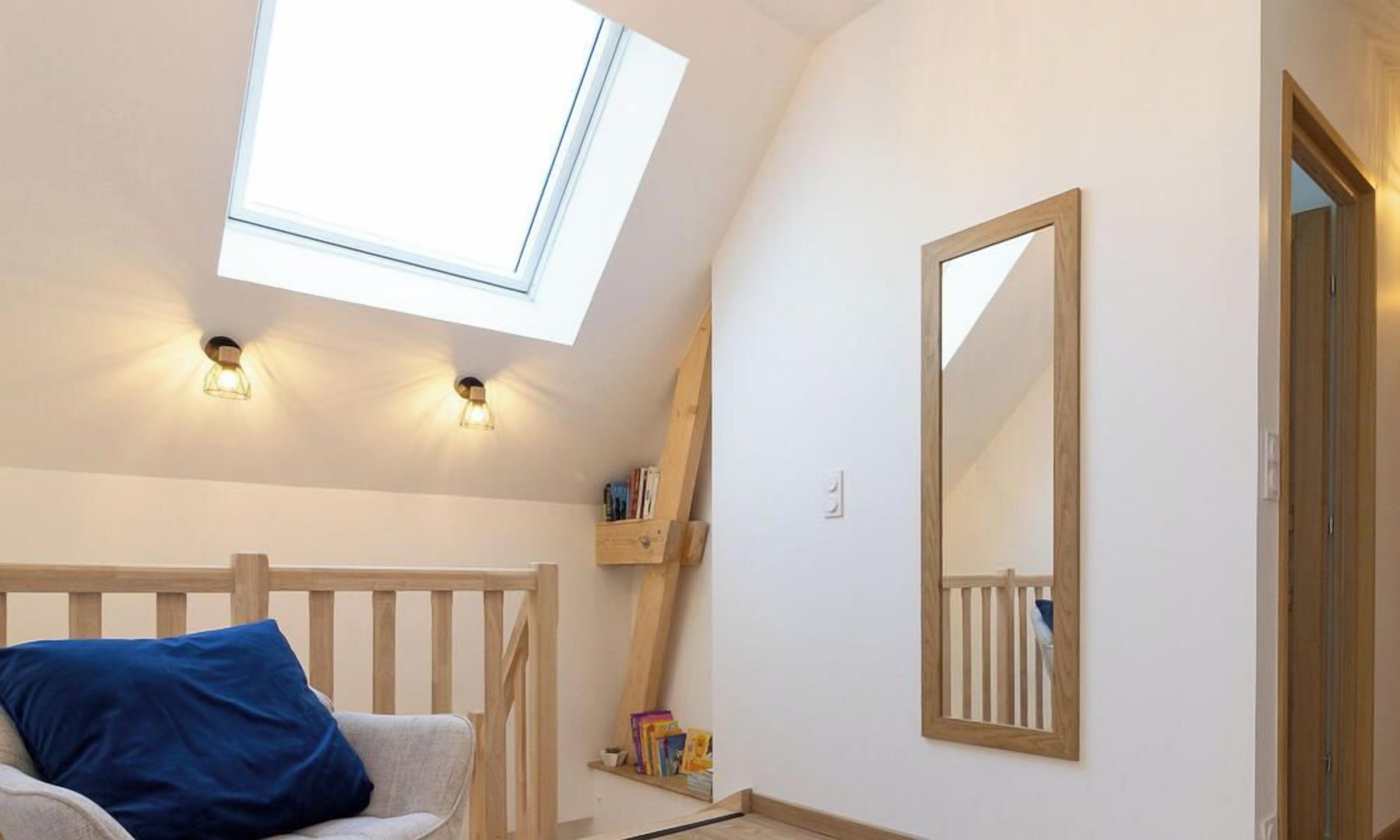 A room with a sloped ceiling, a skylight, a white wall, a wooden bench, a blue cushion, a wooden shelf with books, and a mirror.