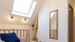 Una habitación con techo inclinado, tragaluz, pared blanca, banco de madera, cojín azul, estante de madera con libros y espejo.