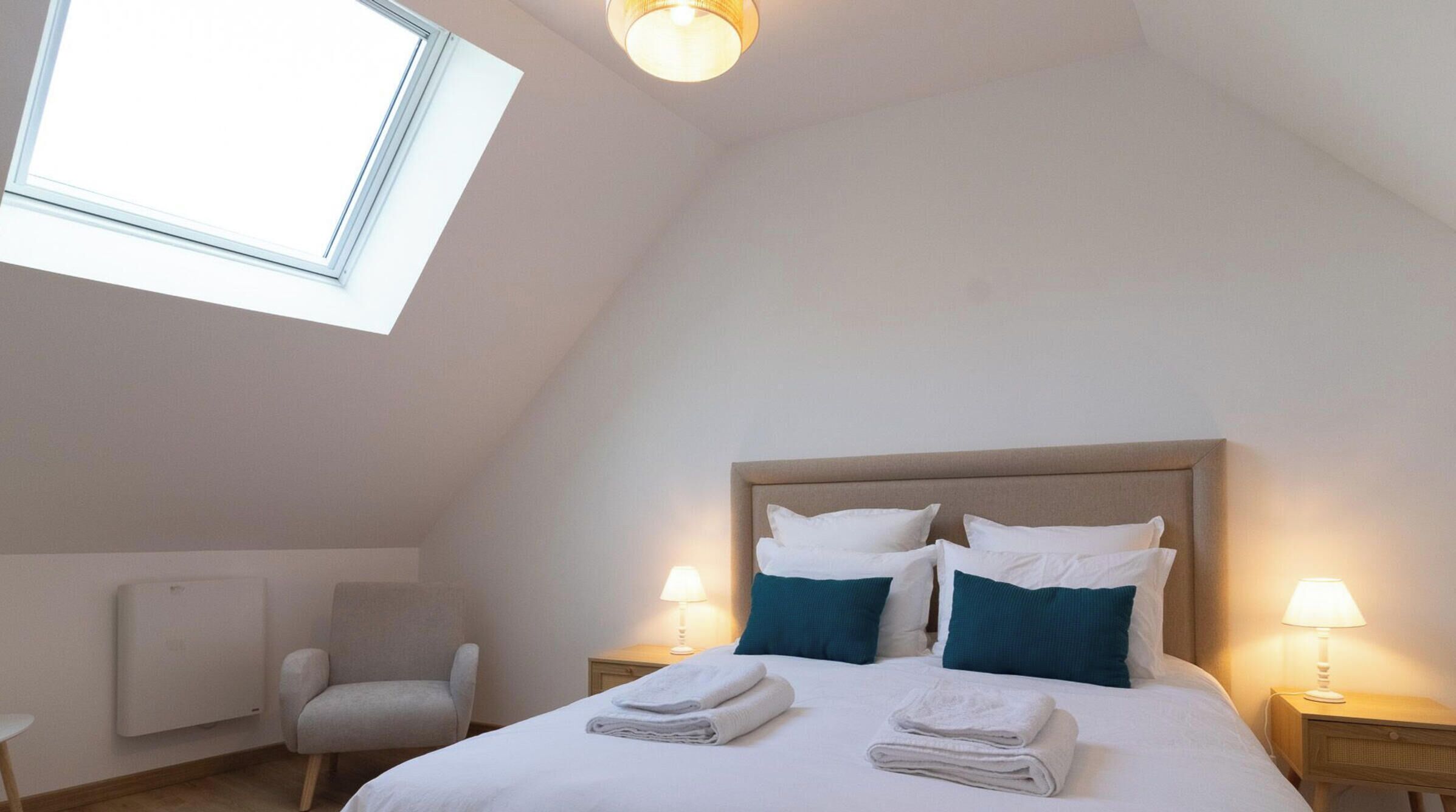 Une chambre avec un plafond en pente, un lit avec une literie blanche et des coussins turquoise, deux lampes de chevet, une chaise et un radiateur.