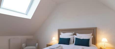 Un dormitorio con techo inclinado, cama con ropa de cama blanca y almohadas color turquesa, dos lámparas de noche, una silla y un radiador.