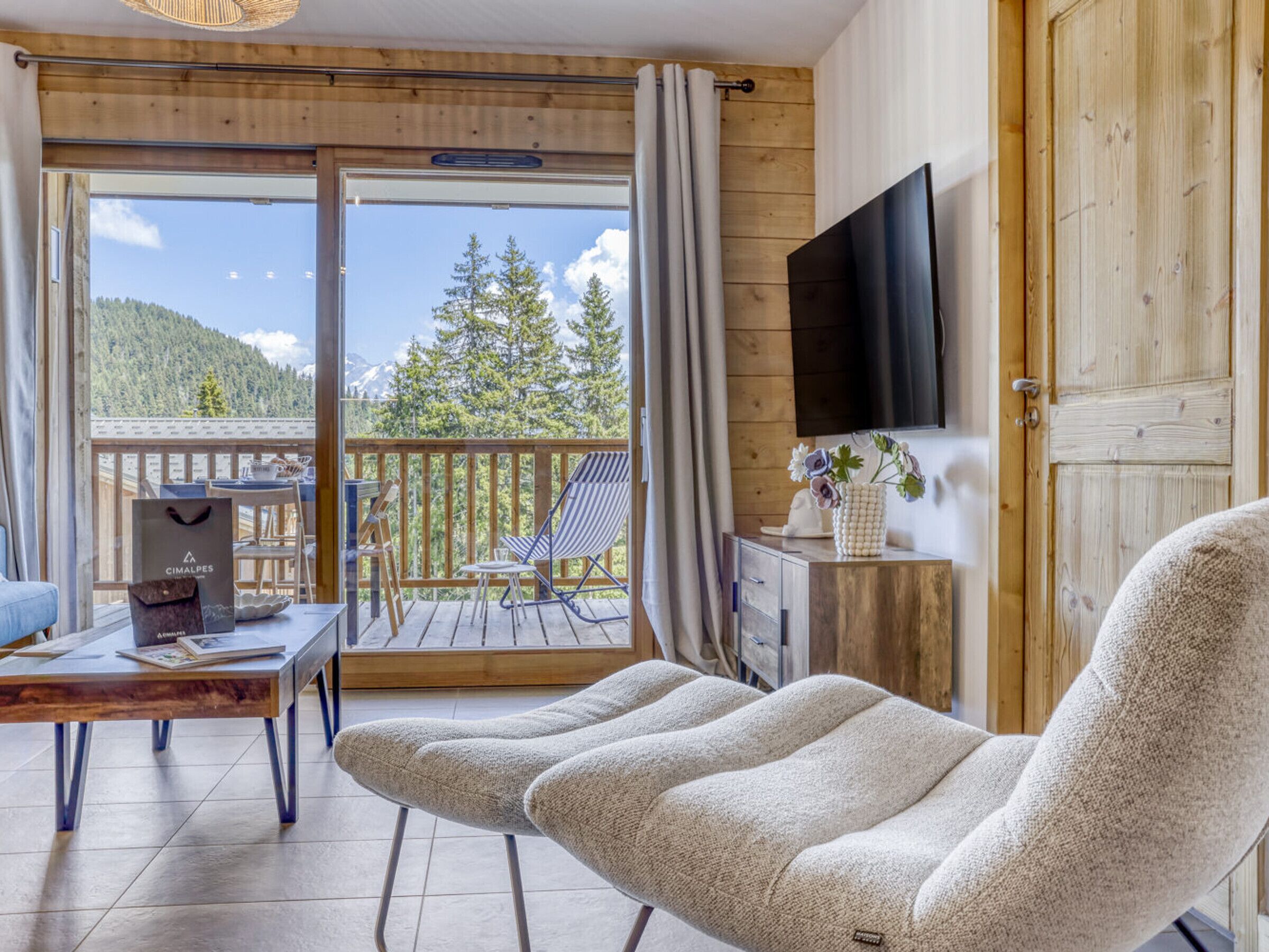 Una habitación con sofá, mesa, televisión y balcón con vistas a árboles y montañas.