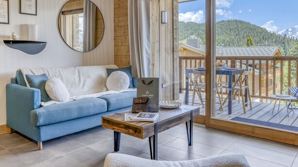 En moderne stue med sofa, sofabord og udsigt til en træterrasse og bjerge.