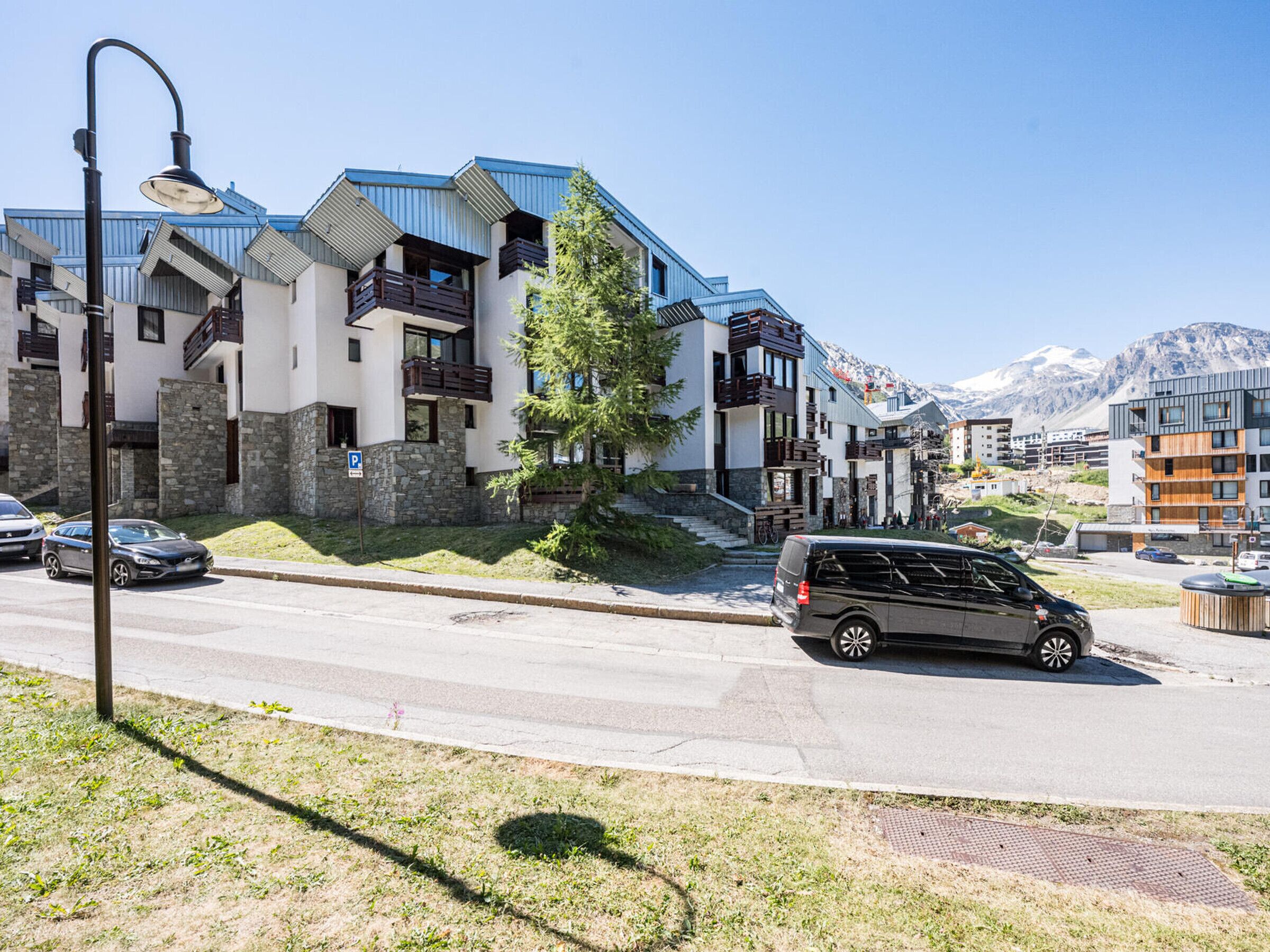 Un moderno edificio de varios pisos con balcones, un área de estacionamiento y montañas al fondo.
