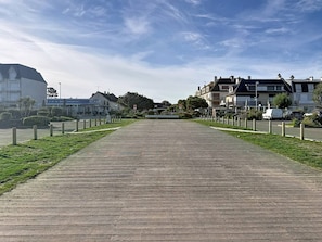 A wide, flat walkway with a wooden surface, flanked by low-rise buildings and greenery. - Apartment with terrace - 5 guests (Merville-Franceville-Plage)