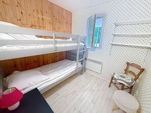 A bunk bed room with a wooden headboard, a window with a view, a radiator, a small table with a checkered tablecloth, and a chair. - Apartment with terrace - 5 guests (Merville-Franceville-Plage)