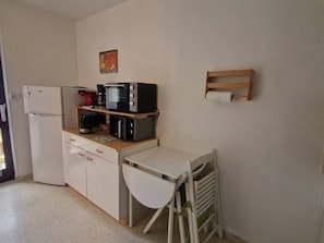 A compact kitchen with a refrigerator, a microwave, and a stove. There is a small table with a chair, and a wooden shelf above the stove. - Apartment with AC, private parking - 4 guests (Balaruc-les-Bains)