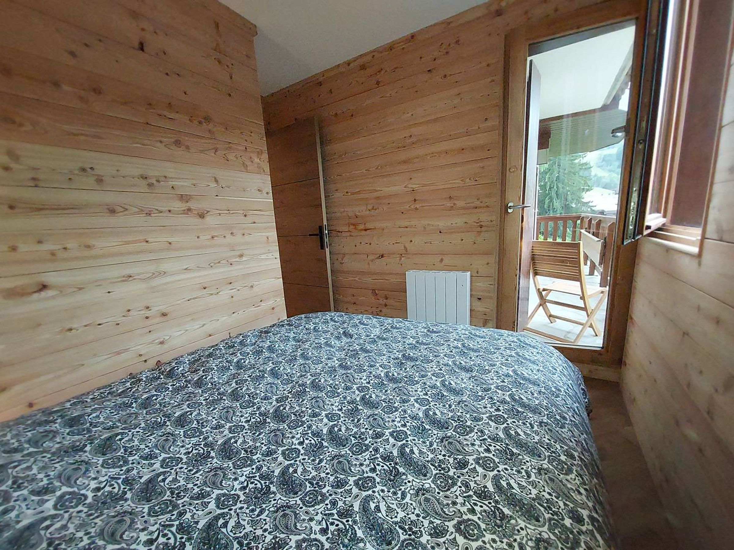 A bedroom with a bed, a radiator, and a sliding door leading to an outdoor area with a chair.