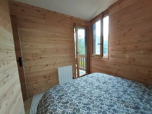 A room with wooden walls, a bed with a patterned bedspread, a radiator, and a window with a view of greenery. - Cozy apartment with panoramic view - 4 guests (Valmorel)