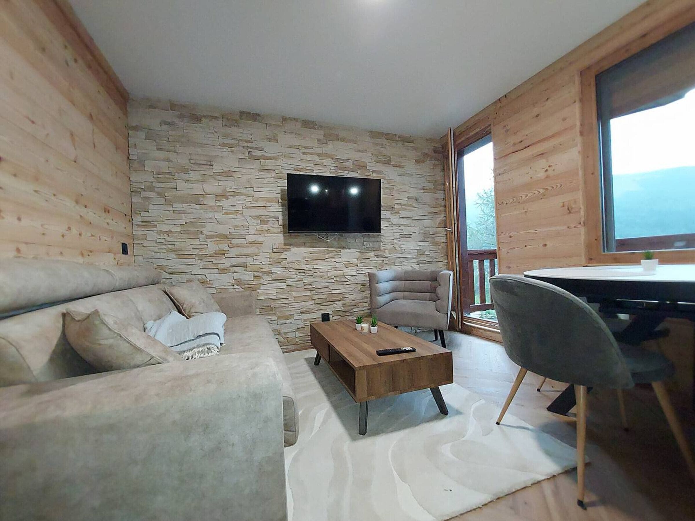 A modern living room with a stone wall, a flat-screen TV, a sofa, a coffee table, and a dining area with a chair.