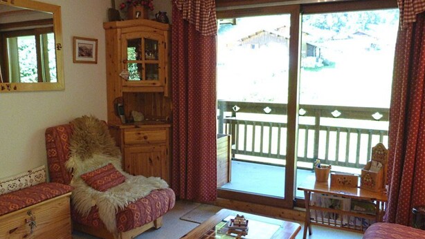 Ein gemütliches Zimmer mit einem Holzkasten, einem gepolsterten Sessel, einem kleinen Tisch und Blick ins Freie durch eine Schiebetür.