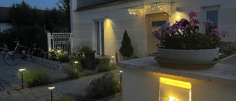 A residential building with a well-lit entrance, a potted plant with flowers, and a number plaque.