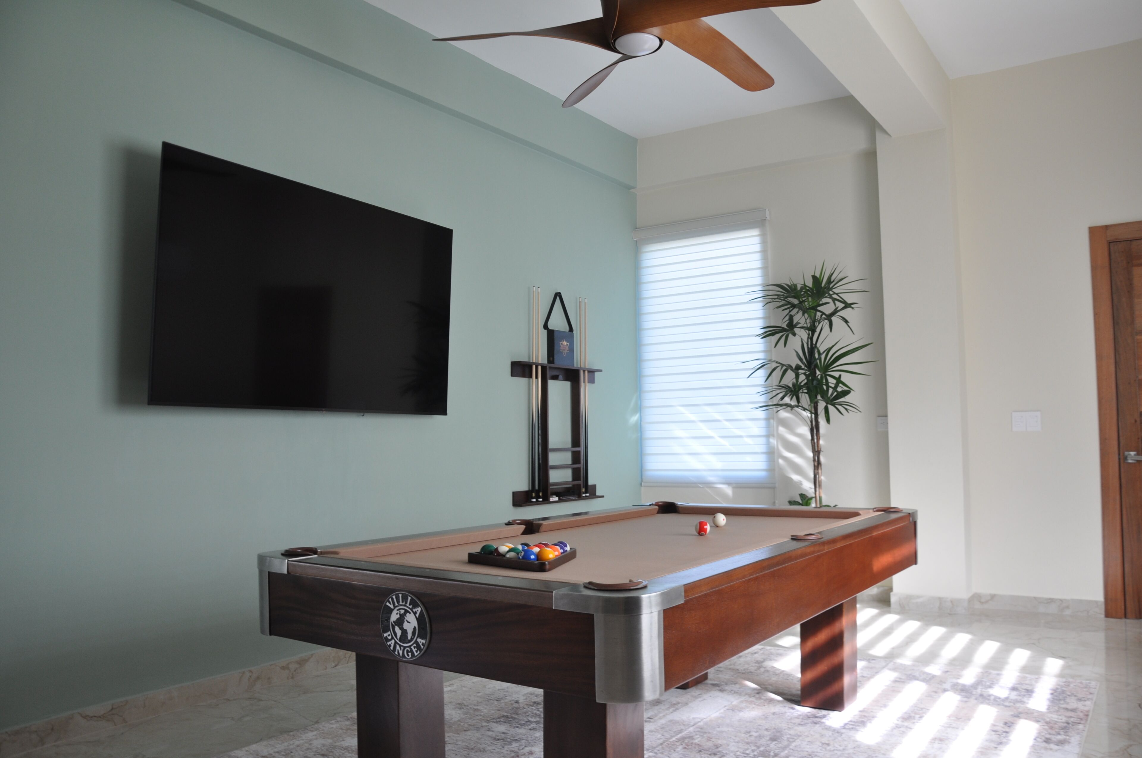 A pool table with a logo, a television mounted on the wall, a ceiling fan, and a potted plant.