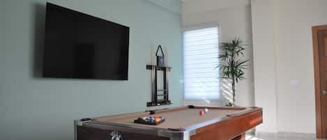 A pool table with a logo, a television mounted on the wall, a ceiling fan, and a potted plant.