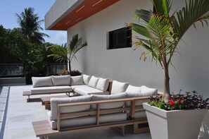 A modern outdoor seating area with a sectional sofa and a wooden table, surrounded by potted plants and a palm tree. - Private 5BD Luxury Villa in Seahorse Ranch w/ Chef, Pool, and Beach Access (Sosúa)