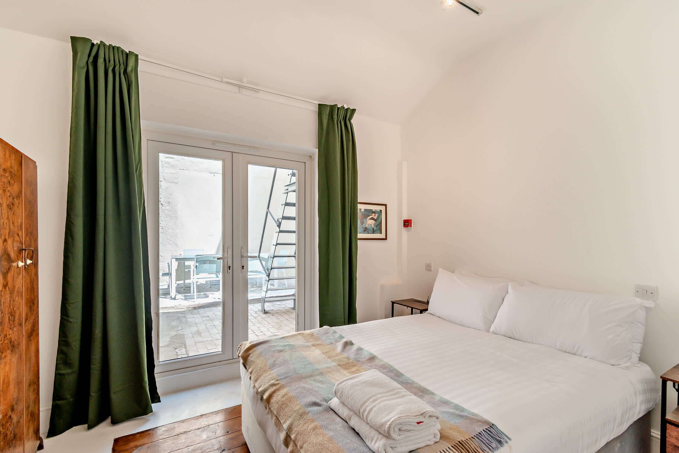 A bedroom with a bed, a wooden wardrobe, a window with curtains, and a small table.