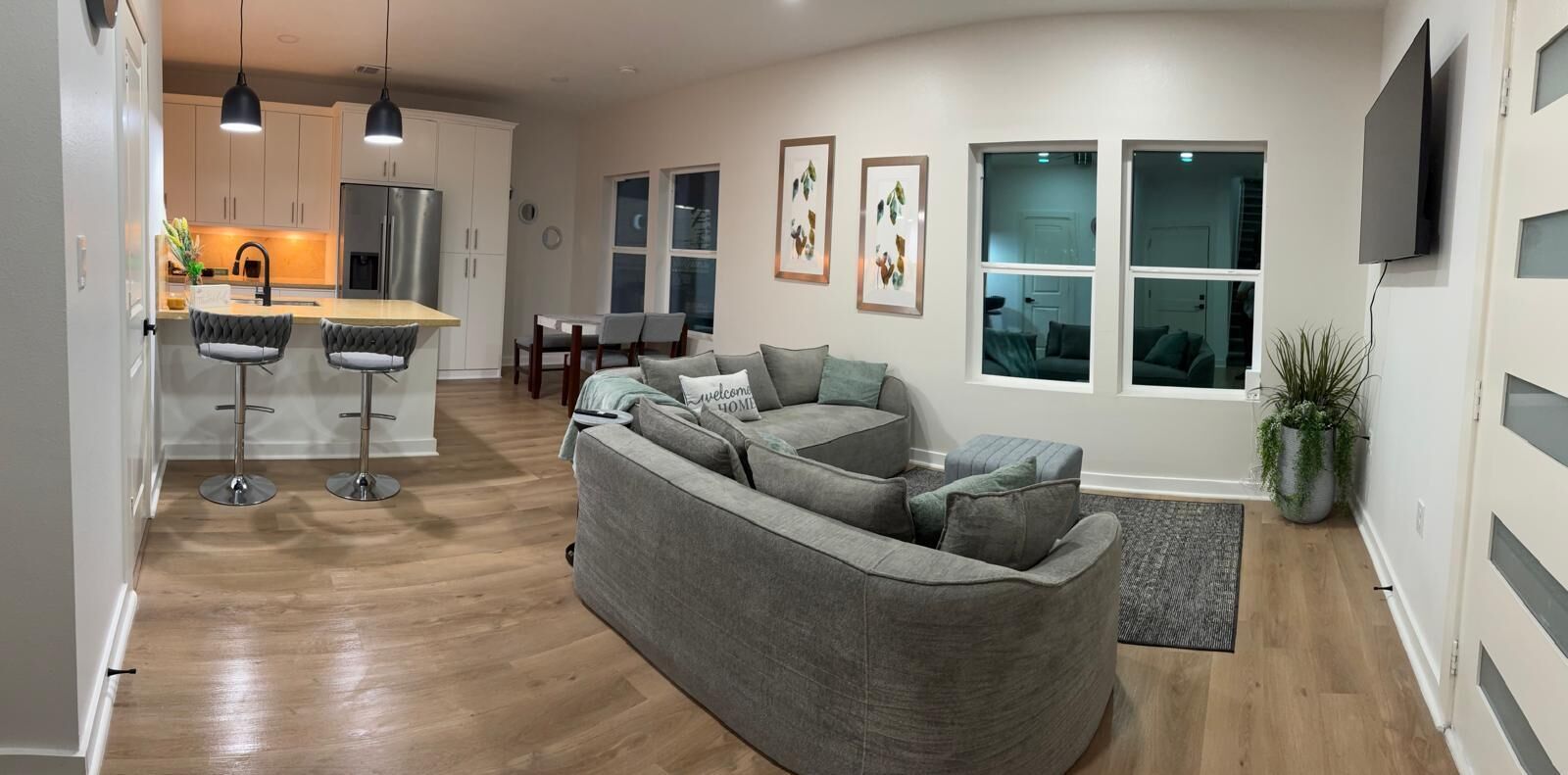 A modern living room with a grey sectional sofa, a dining area with a table and chairs, and a kitchen with white cabinets and a stainless steel refrigerator.