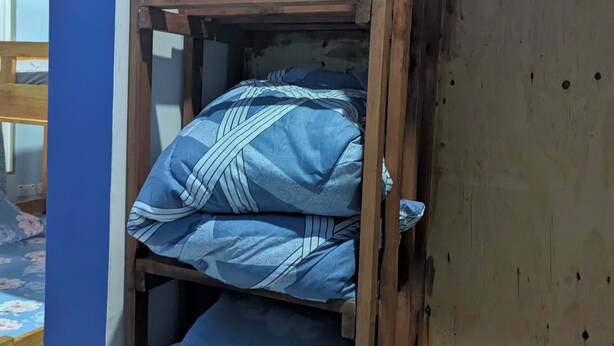 Ein Stockbett mit blauer Bettwäsche und einem floralen Kissen, ein Holz-Kopfstück sowie ein Kleiderschrank im Hintergrund.