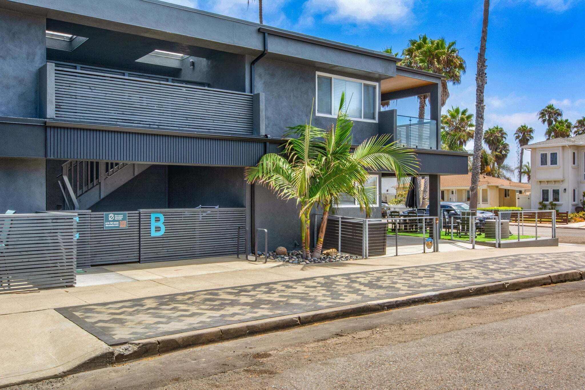 A modern two-story building with a covered outdoor area, a palm tree, and a gated entrance.