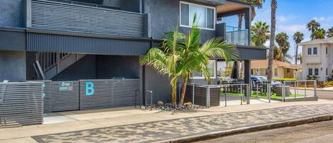 A modern two-story building with a covered outdoor area, a palm tree, and a gated entrance.